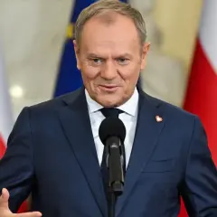 Donald Tusk w garniturze przemawia przy mikrofonie na tle flag Polski i UE.