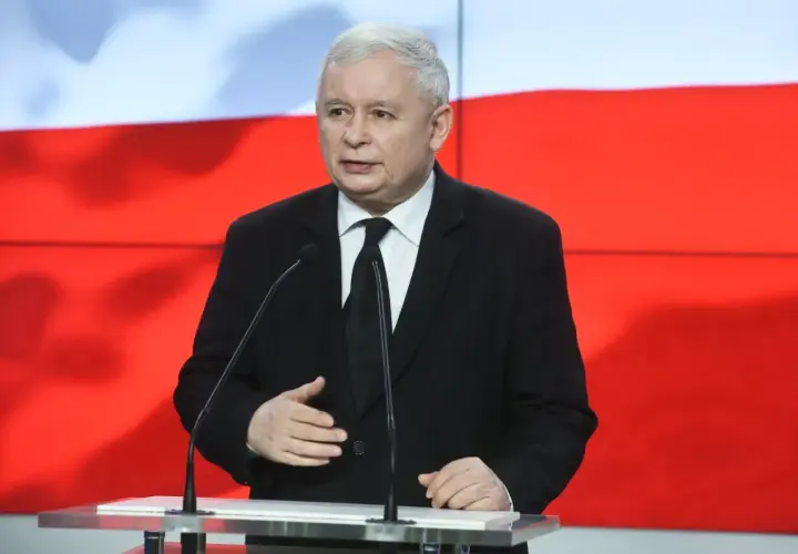 Jarosław Kaczyński w garniturze przemawia przy mównicy na tle flagi Polski.