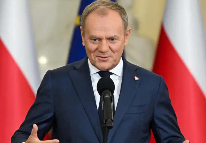 Donald Tusk w garniturze przemawia przy mikrofonie na tle flag Polski i UE.