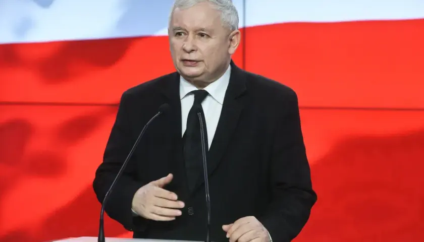 Jarosław Kaczyński w garniturze przemawia przy mównicy na tle flagi Polski.