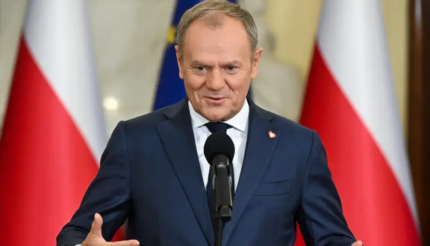 Donald Tusk w garniturze przemawia przy mikrofonie na tle flag Polski i UE.